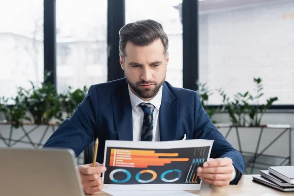 Serious economist looking at charts near blurred laptop and notebooks — Stock Photo