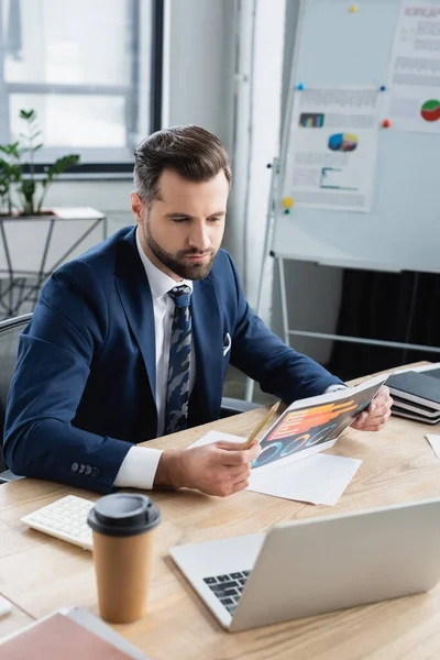 Bärtiger Ökonom betrachtet Analytik in der Nähe von verschwommenem Laptop und Pappbecher — Stockfoto