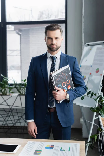 Économiste en costume regardant la caméra tout en se tenant debout avec des graphiques et des dossiers dans le bureau — Photo de stock