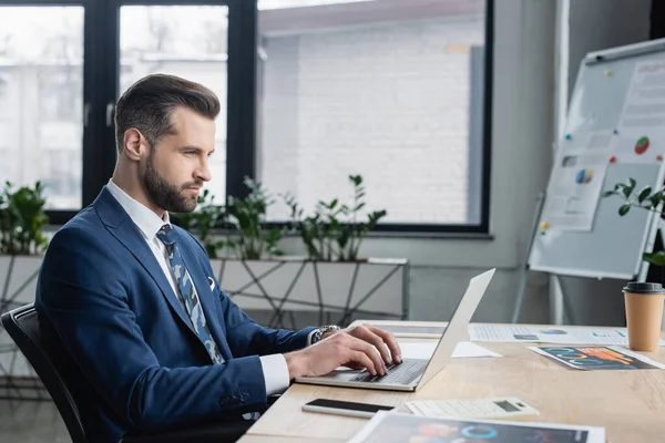 Ökonom tippt auf Laptop in der Nähe von Papieren mit Analysen auf dem Schreibtisch — Stockfoto