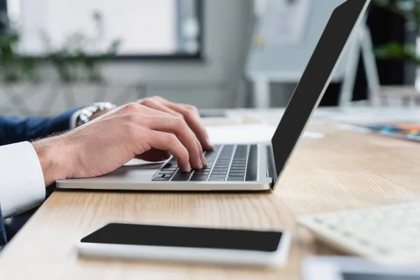 Vista cortada de empresário digitando no laptop perto de smartphone borrado com tela em branco — Fotografia de Stock