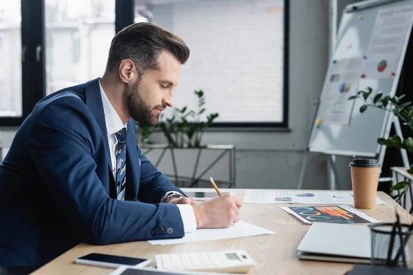 Side view of economist writing near gadgets and graphs on work desk — Stock Photo