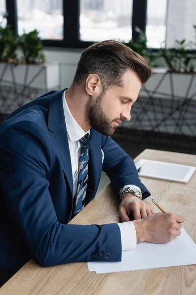 Economista en la escritura de desgaste formal cerca borrosa tableta digital en el escritorio - foto de stock