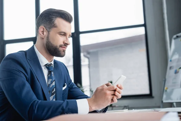 Homme d'affaires dans la messagerie blazer sur smartphone tout en étant assis dans le bureau — Photo de stock