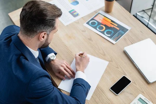 Vista aérea de economista escribiendo cerca de teléfono inteligente con pantalla en blanco y gráficos en el escritorio - foto de stock