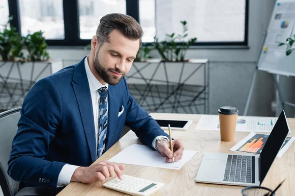 Accountant with pencil using calculator while working near laptop in office — стоковое фото