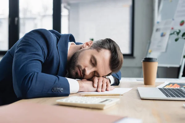 Contable agotado durmiendo cerca de la computadora portátil y café para ir en el escritorio - foto de stock