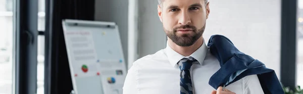 Бизнесмен в белой рубашке, держащий блейзер и смотрящий на камеру в офисе, баннер — стоковое фото