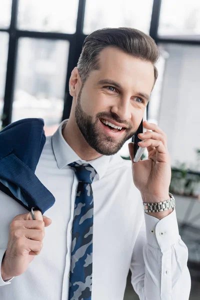 Alegre hombre de negocios hablando en el teléfono móvil mientras sostiene la chaqueta en la oficina — Stock Photo