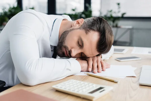 Erschöpfter Ökonom schläft am Arbeitsplatz neben Papieren und verschwommenem Taschenrechner — Stockfoto