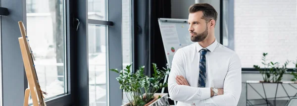 Brunette homme d'affaires en chemise blanche debout avec les bras croisés et regardant loin dans le bureau, bannière — Photo de stock