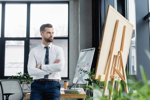 Ökonom im weißen Hemd steht mit verschränkten Armen in der Nähe von Flipcharts und schaut weg — Stockfoto