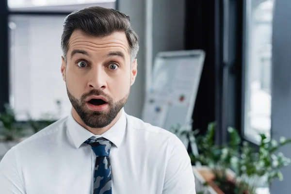 Schockierter Ökonom mit offenem Mund blickt in verschwommenem Büro in die Kamera — Stockfoto