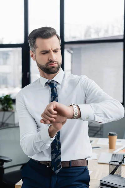 Homme d'affaires sérieux en chemise blanche fronçant les sourcils tout en regardant la montre-bracelet au bureau — Photo de stock