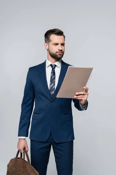 Serious businessman with briefcase reading documents in folder isolated on grey — Stock Photo