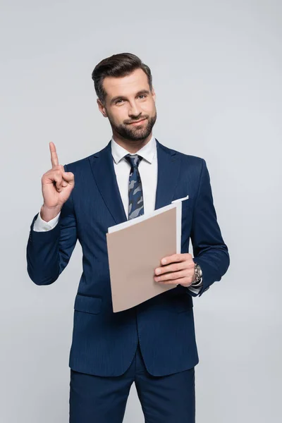 Positivo uomo d'affari tenendo cartella e mostrando idea gesto isolato su grigio — Foto stock