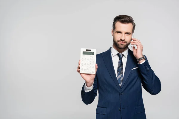 Економіст у синьому костюмі показує калькулятор і розмовляє на мобільному телефоні ізольовано на сірому — стокове фото