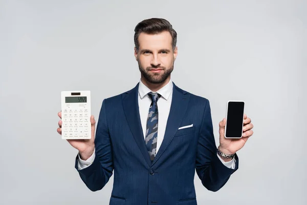 Economista en traje azul que muestra calculadora y teléfono inteligente con pantalla en blanco aislado en gris - foto de stock