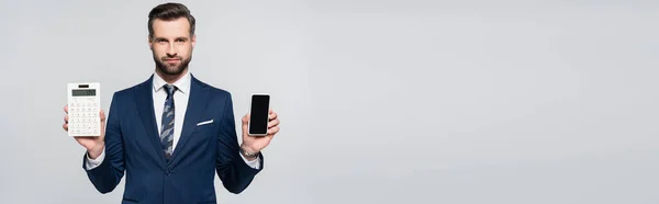 Geschäftsmann im blauen Anzug hält Handy mit leerem Bildschirm und Taschenrechner isoliert auf grau, Banner — Stockfoto