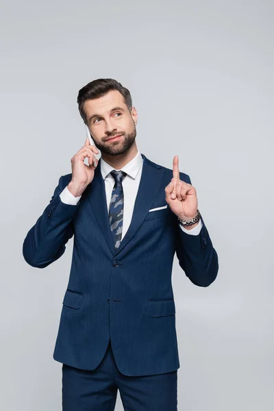 Sonriente hombre de negocios hablando en el teléfono inteligente y mostrando idea signo aislado en gris - foto de stock