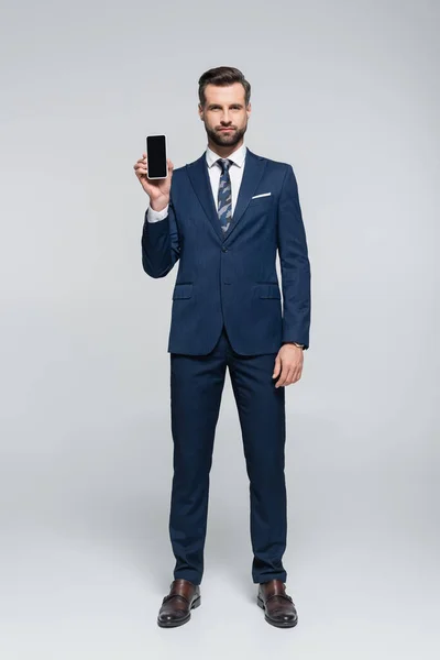 Vue pleine longueur de l'homme d'affaires en costume bleu montrant smartphone avec écran blanc sur gris — Photo de stock