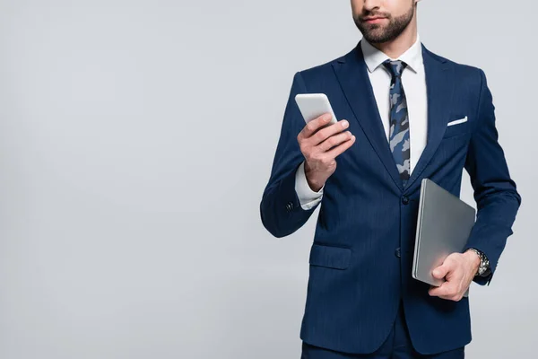 Vista parcial del hombre de negocios con portátil que sostiene el teléfono móvil aislado en gris - foto de stock