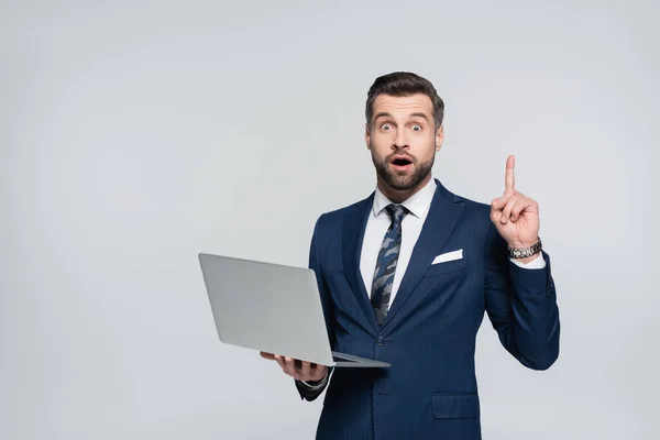 Homme d'affaires étonné tenant ordinateur portable et montrant signe d'idée isolé sur gris — Photo de stock