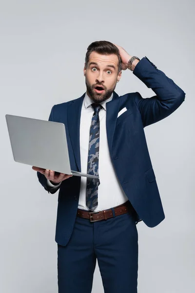 Amazed economist with laptop touching head and looking at camera isolated on grey — стоковое фото