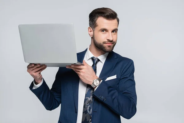 Positive businessman looking at camera while holding laptop isolated on grey — стоковое фото