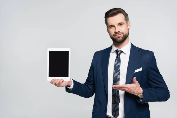 Smiling economist looking at camera and pointing at digital tablet isolated on grey — стоковое фото