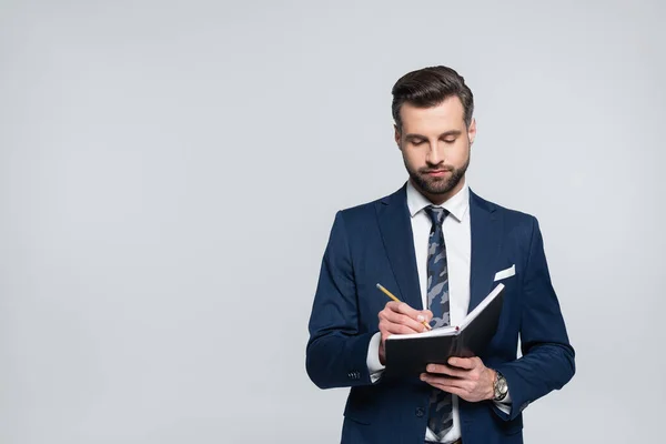 Brunette businessman in blue suit writing in notebook isolated on grey — стоковое фото