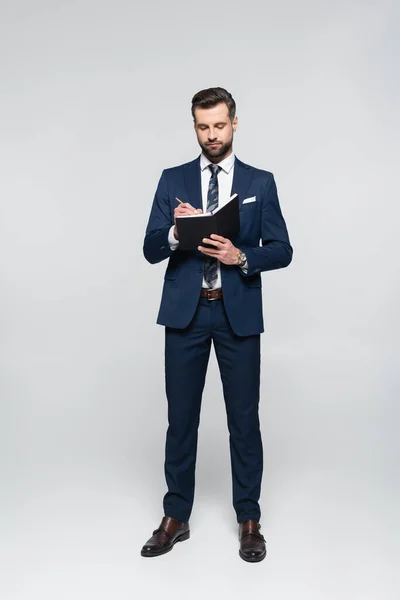 Vue complète de l'économiste barbu en costume écrit dans un cahier sur gris — Photo de stock