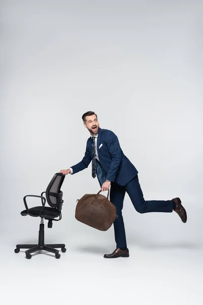 In voller Länge aufgeregter Geschäftsmann mit lederner Aktentasche in der Nähe von Bürostuhl auf grau — Stockfoto