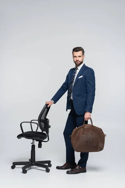 Full length of businessman in blue suit standing with leather briefcase near office chair on grey — стоковое фото