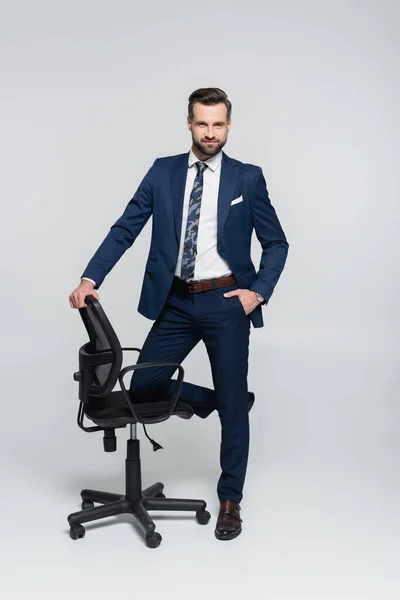 Successful businessman standing with hand in pocket near office chair on grey — стоковое фото