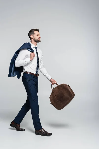 Brünetter Geschäftsmann mit Lederaktentasche und Blazer in voller Länge, der auf grau läuft — Stockfoto