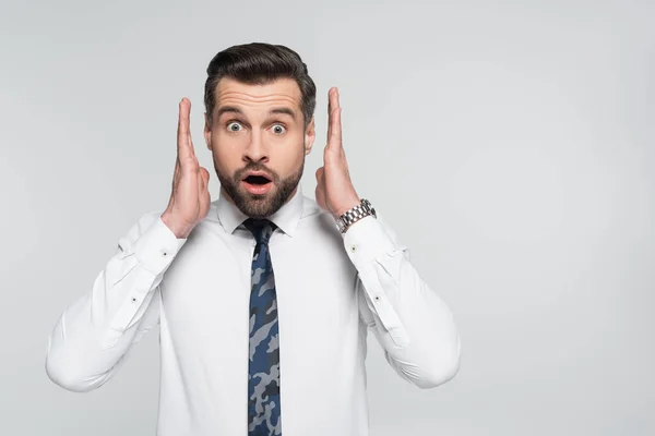 Scioccato uomo d'affari che si tiene per mano vicino al viso e guardando la fotocamera isolata su grigio — Foto stock