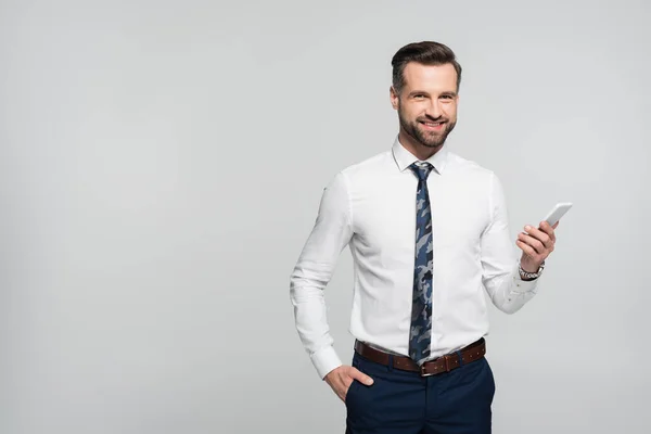 Alegre hombre de negocios de la mano en el bolsillo y el teléfono móvil aislado en gris - foto de stock