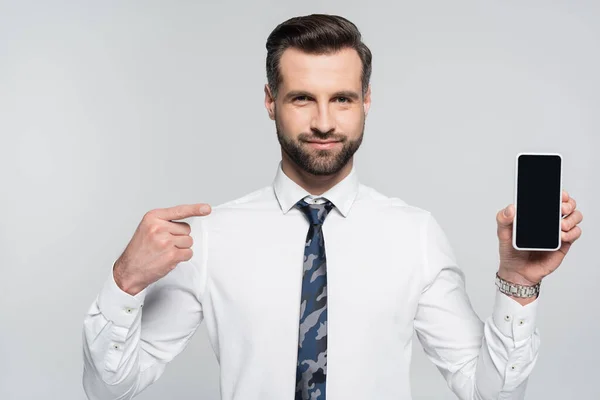 Uomo d'affari sorridente in camicia bianca che punta verso smartphone con schermo bianco isolato su grigio — Foto stock