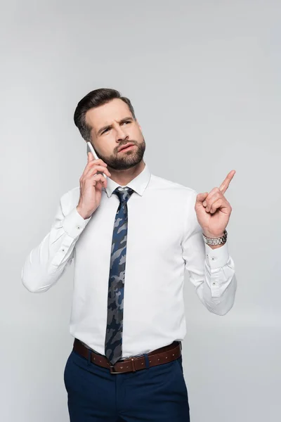 Pensativo hombre de negocios hablando en el teléfono inteligente y señalando con el dedo aislado en gris - foto de stock