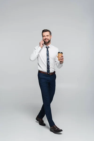 Full length view of happy economist talking on smartphone while walking with paper cup on grey — Stock Photo