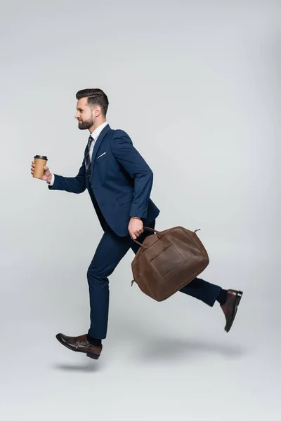 Vista lateral del hombre de negocios en traje azul corriendo con maletín y taza de papel en gris - foto de stock