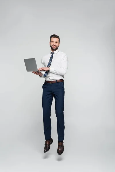 Full length of cheerful economist in formal wear levitating with laptop isolated on grey — стоковое фото