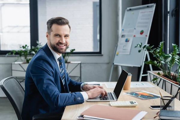 Feliz hombre de negocios mirando a la cámara mientras escribe en el ordenador portátil con pantalla en blanco - foto de stock