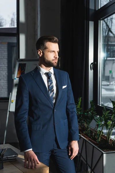 Hombre de negocios serio en traje mirando hacia otro lado mientras está de pie en la oficina - foto de stock