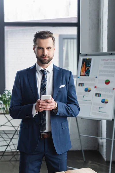 Économiste avec smartphone debout près de tableau à feuilles floues et regardant la caméra — Photo de stock