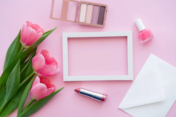 Top view of decorative cosmetics near envelope, frame and blooming tulips on pink — Stock Photo