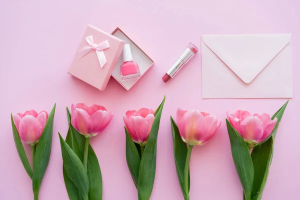 Top view of row with blooming tulips near gift box with nail polish near lipstick and envelope on pink — Stock Photo