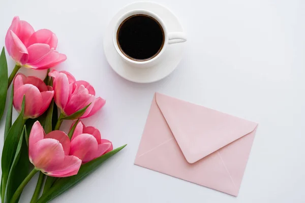 Vista dall'alto di mazzo di tulipani vicino a tazza di caffè e busta rosa su bianco — Foto stock