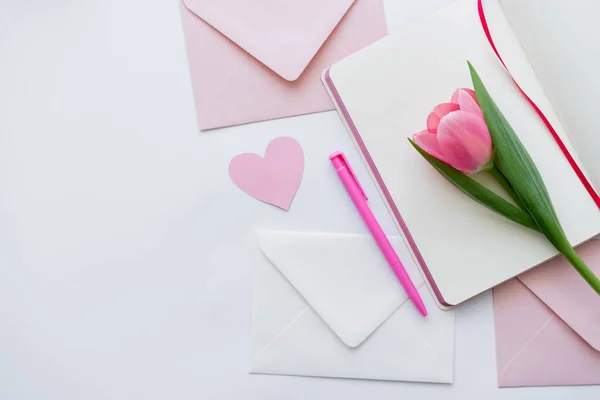 Top view of pink tulip on notebook near envelopes and paper heart on white — Stock Photo